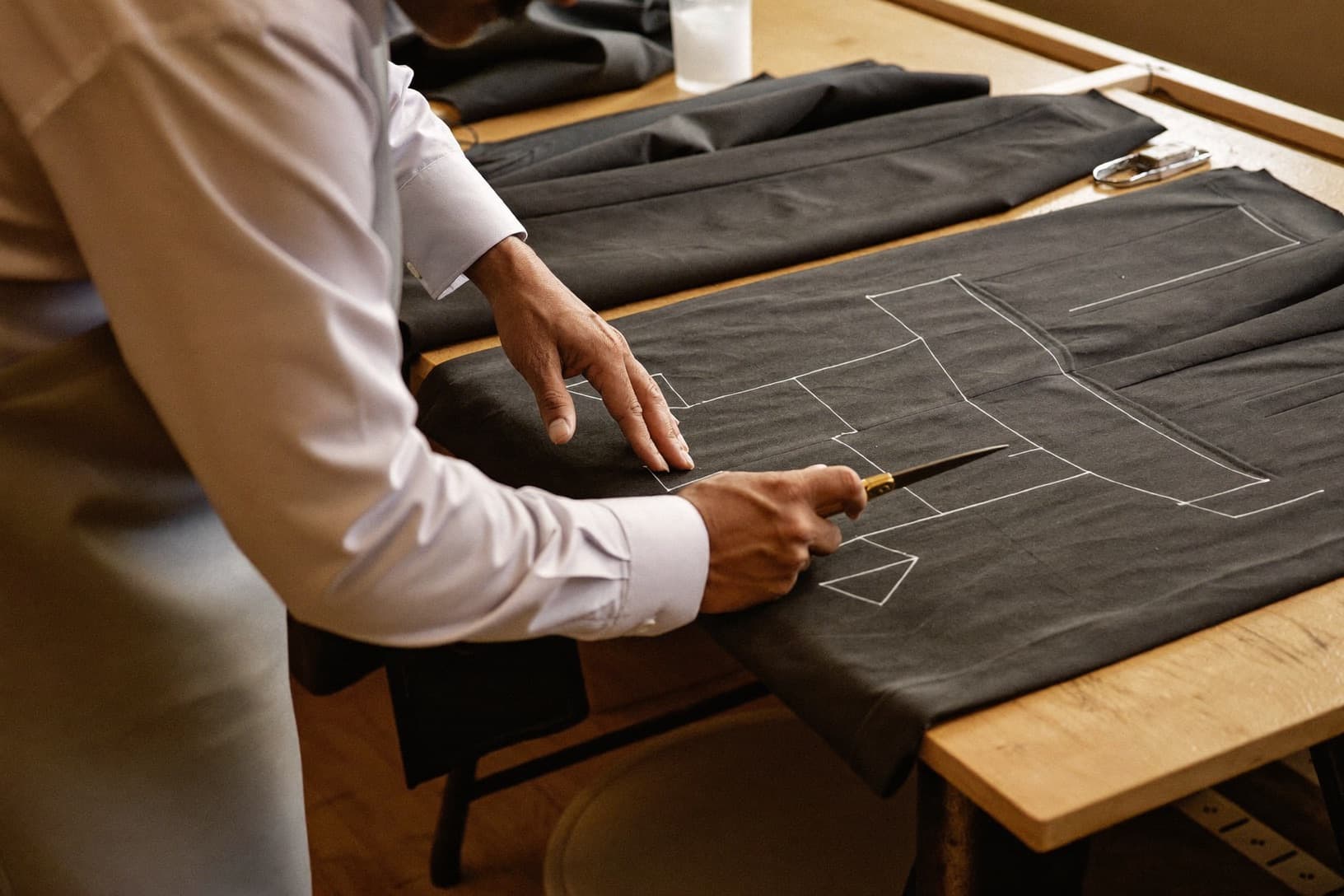 A master tailor draws chalk lines across dark suiting cloth on a cutting table.